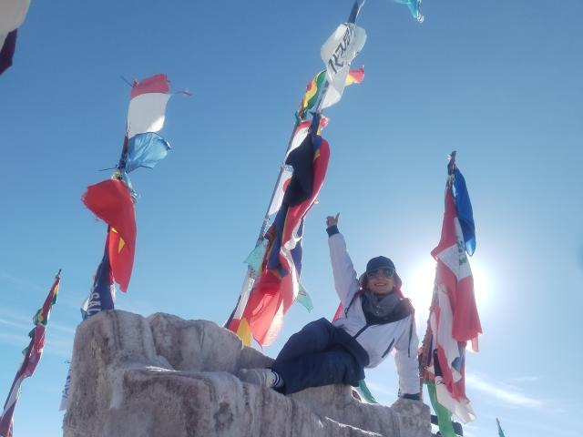 AmyStreetwise69 with her ass on a stone, wearing a blue jacket and gray beanie, raising a finger against a bright blue sky with colorful flags. Sun in the background.