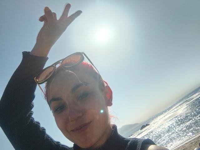 Amy, capelli rossi, occhiali da sole in testa, maglietta scura, sorridente, sole splendente, ammicca con la mano destra alzata, cielo luminoso, riflesso del sole, spiaggia, acqua scintillante, atmosfera rilassata..