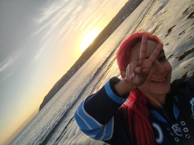 AmyStreetwise69, con cabello rojo, haciendo un gesto de paz, sonriendo, vestida con un traje de neopreno azul y negro, de pie junto a una superficie acuática iluminada por el sol y reflectante, mostrando su linda cara..