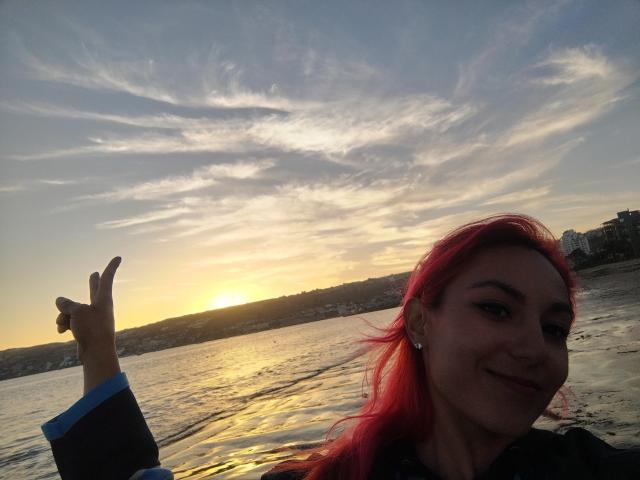 AmyStreetwise69 with red hair, smiling, holding up a peace sign, wearing a black shirt, standing by a sunsetting water, reflecting golden light, showing her nose ring and earrings.