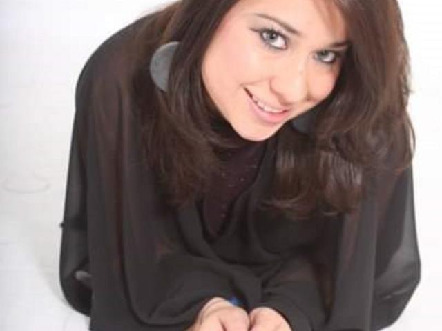 EvaClarks with medium brown hair, black sheer top, and grey earrings, smiling and looking up with dark eyes, her small tits barely visible under the loose top.