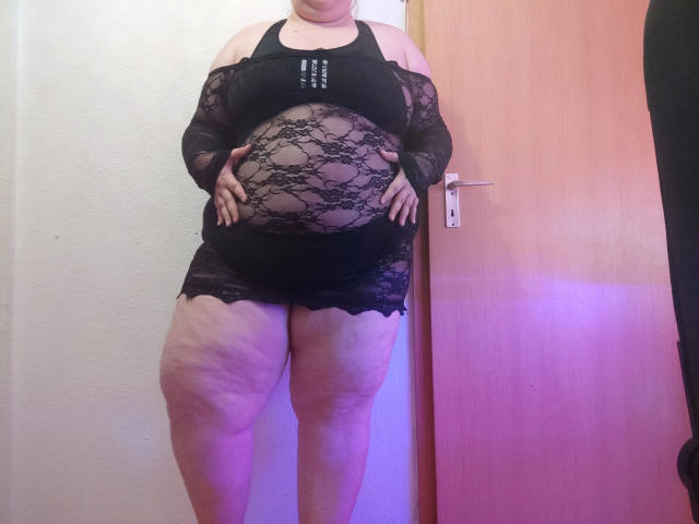 ChubbyBubby-hot, obese with large belly, wearing black lace top and skirt, standing against wooden door, hands on belly, showing fat thighs and stretch marks.