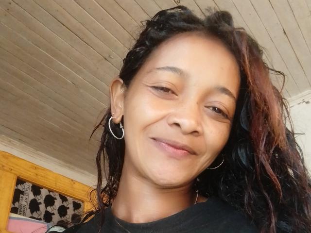 NayaHot has brown skin, curly black hair with red highlights, small hoop earrings, and a subtle smile. She's wearing a black shirt, looking slightly to the side.