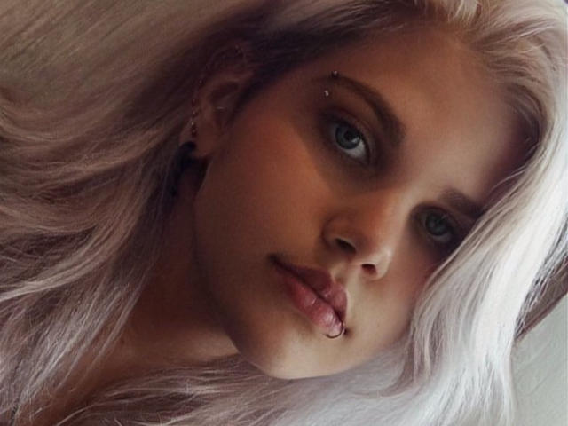 JoannLowe with platinum blonde hair, blue eyes, and a lip piercing, looking at the camera with slightly pouted lips, showing a small amount of her tits.