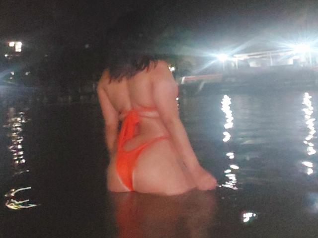 LauraRichis in a red thong bikini, back to the camera, wading in dark water with light reflections, showing her ass and back, with wet, dark brown hair.