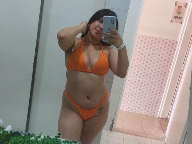 LauraRichis in an orange bikini, showing her tits and pussy in a mirror selfie, with her hand in her hair, a tattoo on her right hip, and a smile.