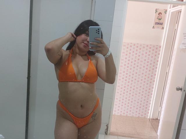 LauraRichis in an orange bikini, showing her tits and pussy in a mirror selfie, with a hand in her hair, a white wristband, and a tattoo on her right thigh.