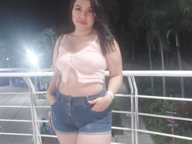 LauraRichis with black hair, pink tied crop top showing her tits, and blue denim shorts with hands in pockets, standing on a balcony at night.