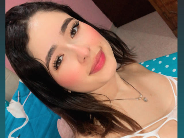 ElinaLeducc ha i capelli scuri, un leggero rossore sulle guance e rossetto rosa. Indossa una maglietta bianca con una collana, e il suo seno è leggermente visibile. Sorride con un'espressione carina..