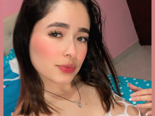 ElinaLeducc has light skin, dark brown hair, and wears a white top. Her pink blushed cheeks and red lips stand out, with a necklace featuring a small pendant.