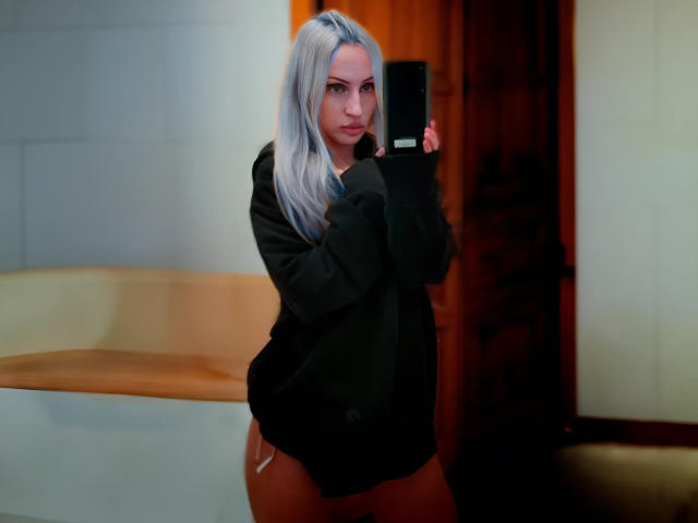LindaFlirt with silver hair, black sweater, and no pants, holding a camera, showing her ass, looking at the camera with a slight pout.