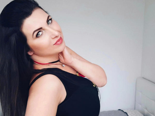ReifeMirabelle-hot with black hair, green eyes, and pink lipstick, wearing a black top and red necklace, touching her neck, smiling softly. Boobs slightly visible, fair skin.