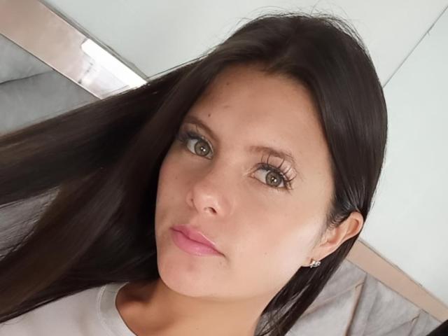 VictoriaWaynes has long dark brown hair, brown eyes, and pink lipstick. She's wearing a white shirt with small earrings and looking at the camera with a slight pout.