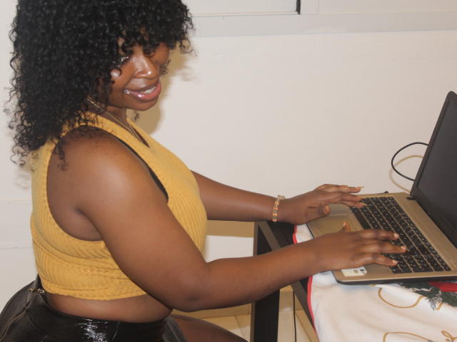 Annykaa, a dark-skinned woman with curly black hair, is typing on a laptop. She's wearing a yellow ribbed crop top and black shiny pants.