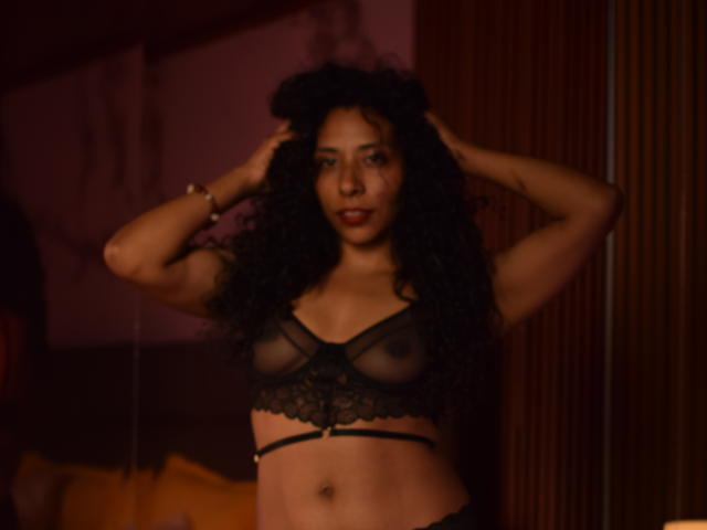 JanEvans with curly black hair, wearing sheer black lace bra, hands in hair, standing, dark skin, small breasts, visible nipples, looking at camera, dim lighting, wooden background.
