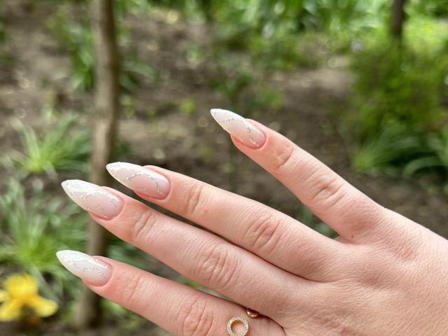 SugarrX's hand with long, pointed, white-nail acrylics, pinkish-nude base, small glitter accents, and a gold ring on the index finger. Background is blurry greenery.