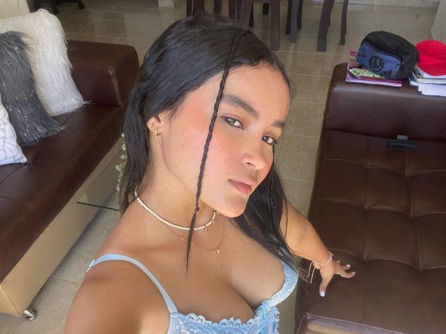 MiiaPalmer, avec de longs cheveux noirs, une peau brun clair, portant un haut en dentelle bleu qui révèle ses petites seins, et un collier en or, se prend en photo dans un salon..