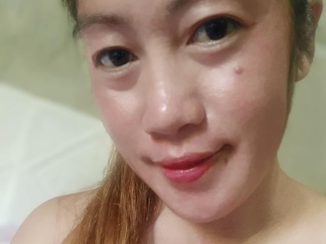 Xenia69's face with light skin, brown eyes, and a small mole on the right cheek, smiling slightly with pink lipstick, and auburn hair tied back.