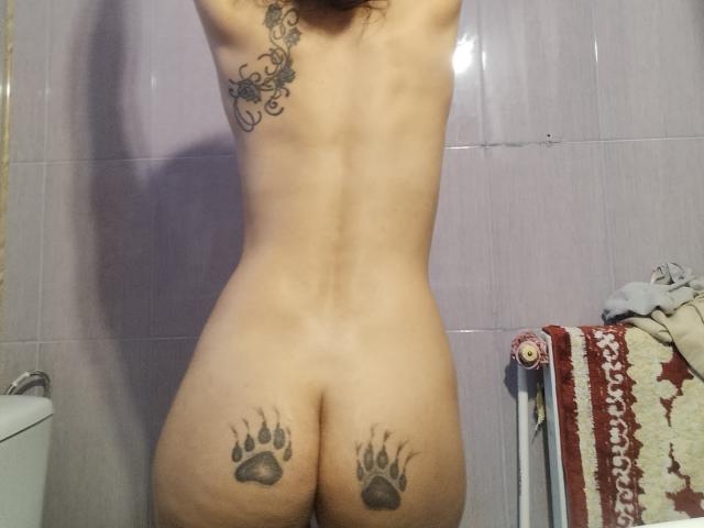 NickySweetX's nude back, arms raised, showing tattoos on lower back and butt, paw prints, and a floral design on left side. She's standing in a bathroom.