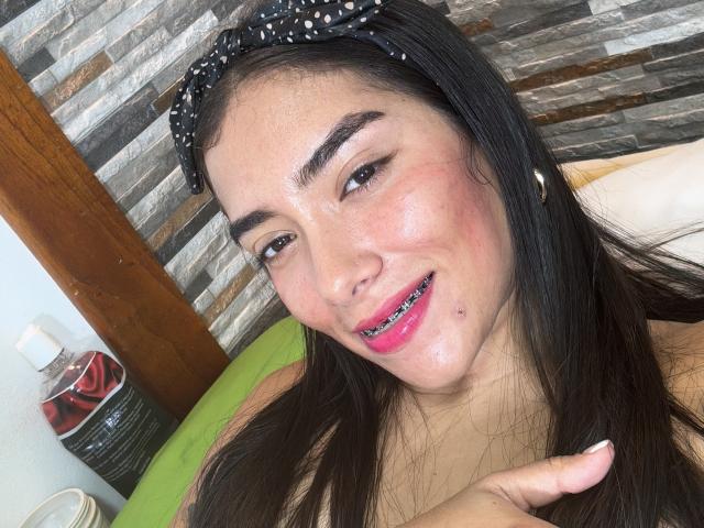 SharonKlart with shiny face, pink lipstick, black hair in polka dot headband, smiling, pointing, small pimple on cheek, brown eyes, and wearing a yellow top.