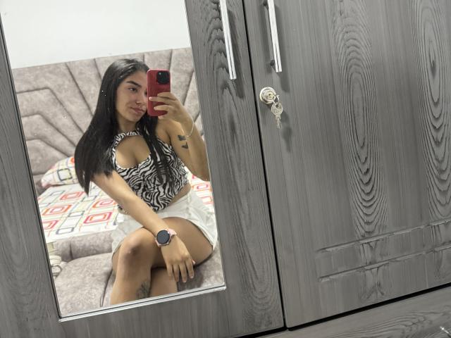 SharonKlart with long black hair, wearing a zebra-pattern halter top and white skirt, sitting on a bed, taking a mirror selfie, showing her tits and a tattoo.