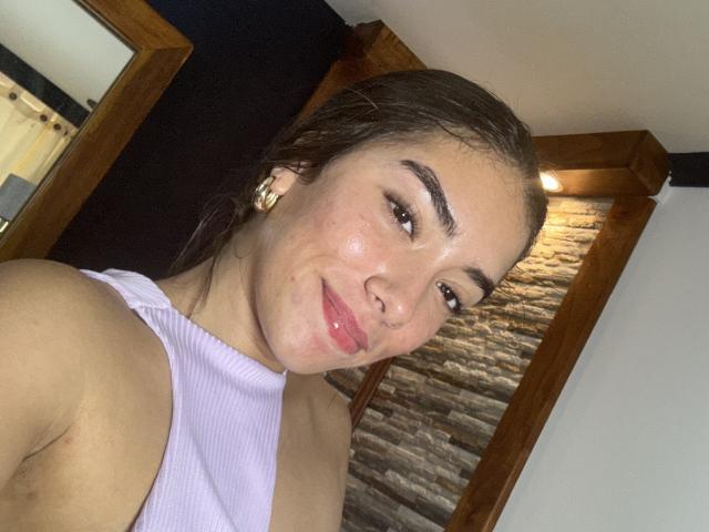 SharonKlart, com lábios rosados e brilhantes, sorrindo levemente, vestindo uma regata roxa, mostrando seu ombro nu e cabelo castanho, tem um rosto bonito e pequenos brincos de ouro..