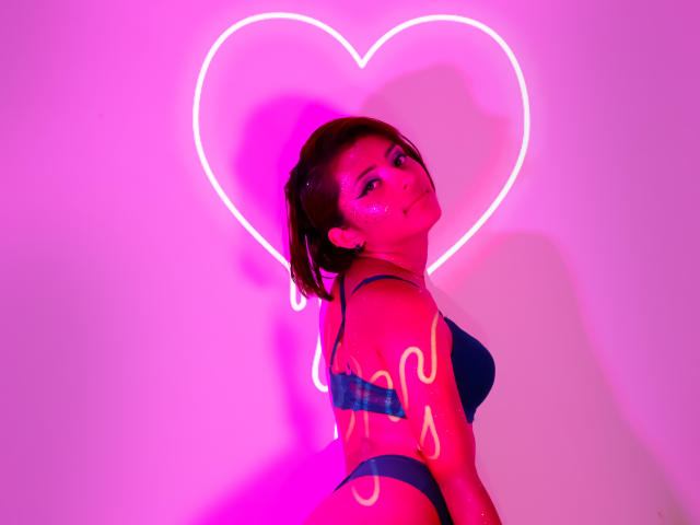 LizBonnet with pink neon heart background, dark blue bra and panties, short brown hair, glowing pink tattoo-like lines on arm, looking back with seductive smile, pink and purple lighting.