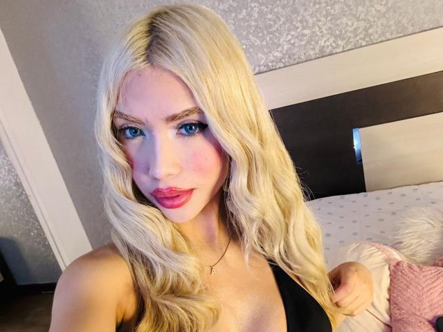 ChristieGarci with long blonde hair, blue eyes, pink cheeks, and red lipstick, wearing a black top, showing her tits, in a bedroom with a bed and pillows.