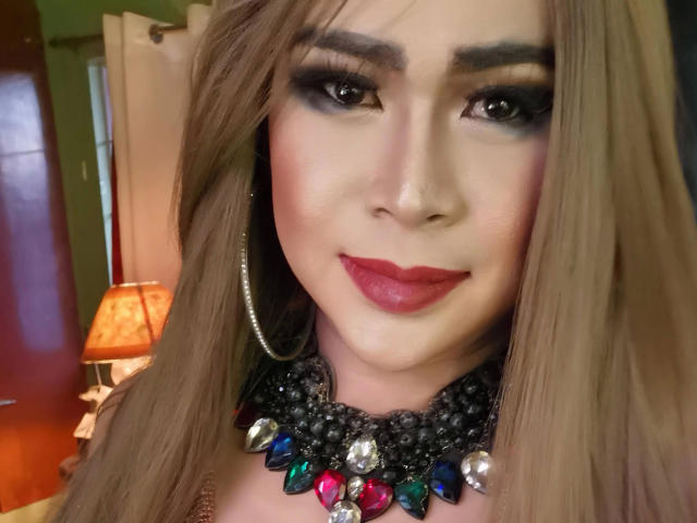 Transsweetdenise-hot with long brown hair, red lipstick, black eye makeup, silver nose ring, and a colorful, jeweled necklace, smiling at the camera. Boobs not visible, focus on face.