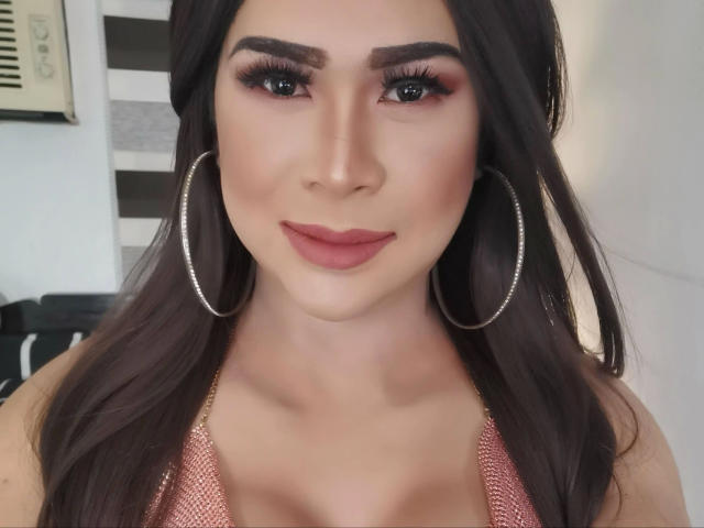 transsweetdenise-hot with long dark hair, wearing large hoop earrings, pinkish-red lipstick, and a textured pink top, showing her tits slightly in the frame, smiling at the camera.