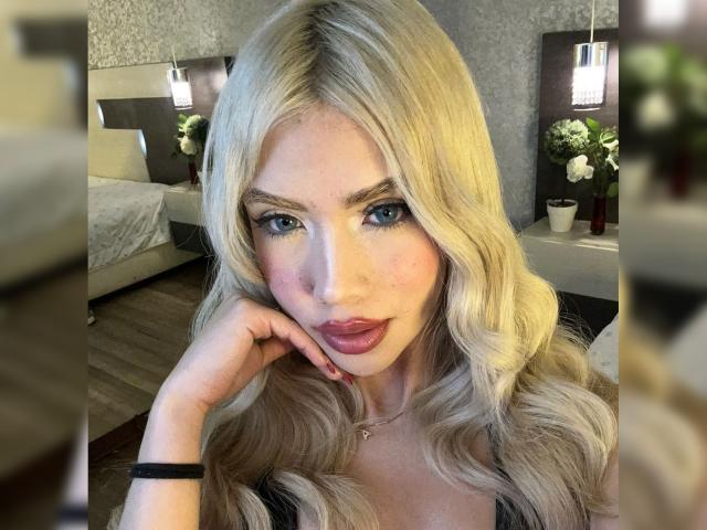 ChristieGarci has blonde wavy hair, blue eyes, and full pink lips. She's touching her cheek with a black bracelet on her wrist, looking at the camera.