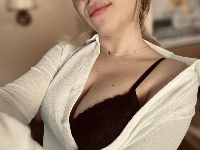 SugarrX with light skin, blonde hair, and a small necklace, wearing an open white shirt revealing black lace bra, showcasing her tits. Soft lighting, slight smile.