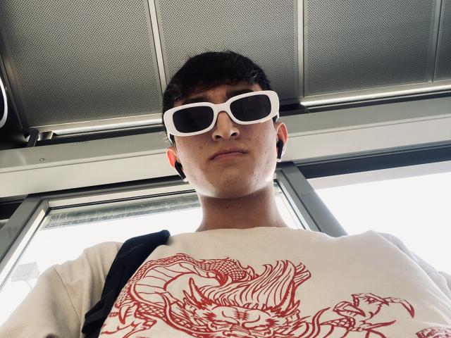 JaySon wearing white sunglasses, short dark hair, white shirt with red dragon design, black strap, looking down at camera, inside bus, ceiling above.