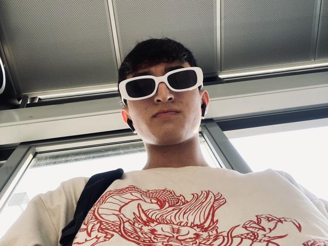 JaySon with white sunglasses, short dark hair, white shirt with red dragon graphic, black shoulder strap, looking down, neutral expression, inside bus, overhead metal panels visible.