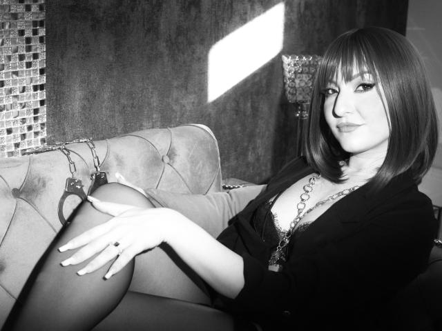 GoddessAlma with bob haircut, black blouse, chain necklace, and stockings, sitting on couch with cuffs on her thigh, looking seductively at camera. Black-and-white photo.