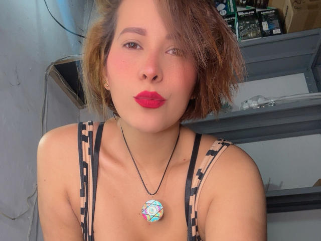 SaritaVelez with short brown hair, red lipstick, and a geometric pendant, wearing a black and beige patterned top, puckering her lips. Boobs visible, light skin, slight blush.