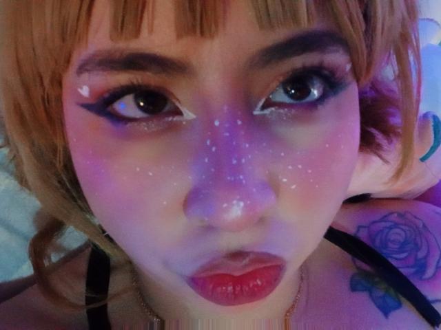 Yuri69s Gesicht mit violettem und rosa Glitzer auf der Nase, roten Lippen, schwarzem Eyeliner und Tattoos an der Schulter, die in die Kamera mit ihren großen, glänzenden Augen blickt..