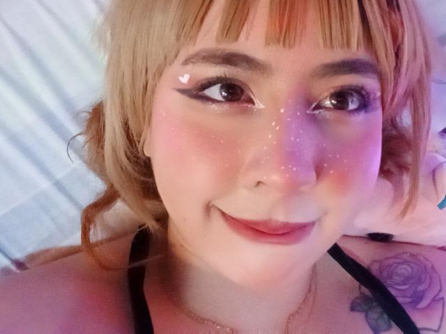 Yuri69 mit blonden Haaren, Eyeliner und Glitzer auf der Wange, lächelt in die Kamera. Sie hat ein Tattoo an ihrer rechten Schulter und trägt ein schwarzes Oberteil..