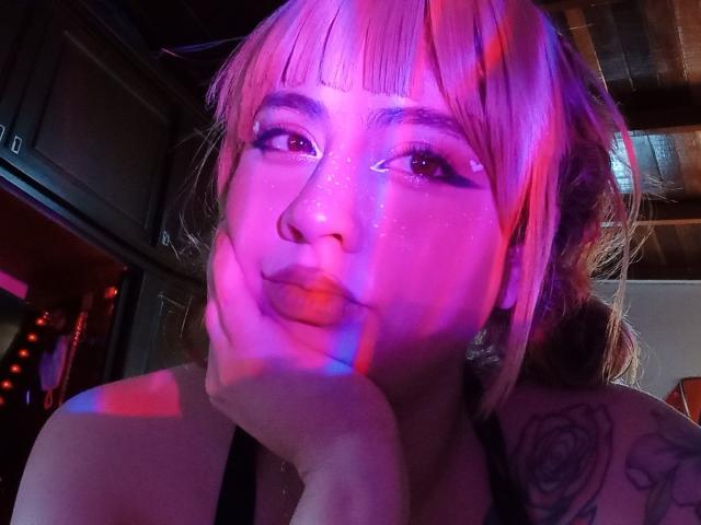 Yuri69 tiene reflejos de luces de neón rosadas y moradas en su rostro, con flequillo, pecas y un hombro tatuado, con el mentón apoyado en la mano, mirando hacia arriba..