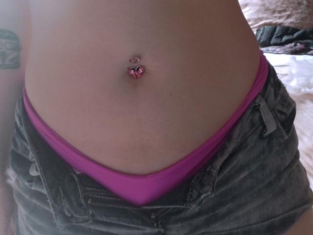 Yuri69 mostra la parte superiore del suo addome con un piercing all'ombelico a forma di cuore rosa, indossa pantaloncini grigi sbottonati e biancheria intima rosa, rivelando un tatuaggio sul lato sinistro..