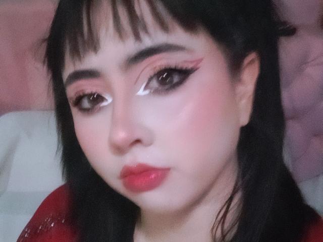 Yuri69, avec une peau pâle, des cheveux noirs, un eye-liner rose et du rouge à lèvres rouge, regarde vers le bas avec un léger sourire, portant un haut rouge, dans un autoportrait légèrement flou..