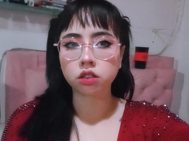Yuri69, avec des couettes noires, des lunettes roses, du rouge à lèvres rouge et un haut rouge pailleté, regarde directement l'appareil photo. Ses fesses ne sont pas visibles. Ses seins sont petits..