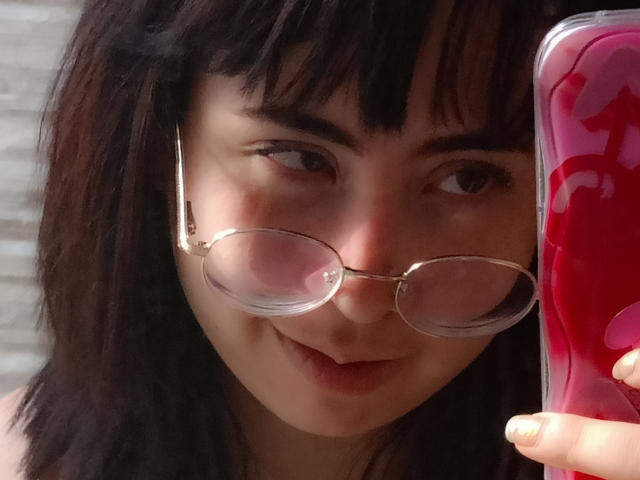 Yuri69 con capelli castano scuro, che indossa occhiali, sorride leggermente, tenendo un telefono rosa con motivi rossi, primo piano, che mostra il naso e le labbra. Il seno non è visibile, l'attenzione è sul viso..