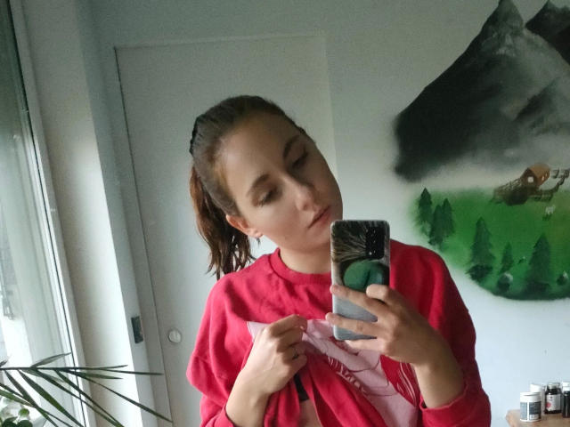 NettePaloma-hot with brown ponytail, red sweatshirt, taking selfie in mirror with phone, small tits, slight pout, white door, green wall art, window, plant, and cans in background.