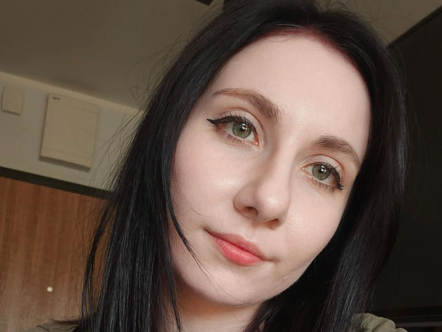 NettePaloma-hot has pale skin, green eyes with black eyeliner, black straight hair, and pink lips. Her nose is small and she's smiling slightly. Her tits are not visible.