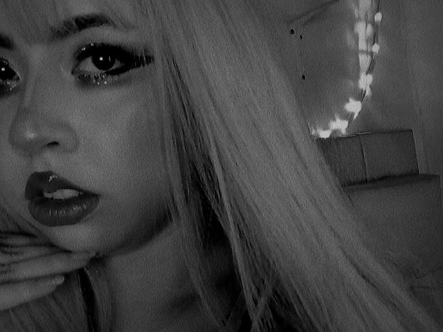 Yuri69 hat langes, glattes blondes Haar, dunklen Eyeliner und glänzlichen Lippenstift. Ihre Hand ist in der Nähe ihres Mundes. Schwarz-weiß-Foto. Ihre Augen sind leicht geöffnet und blicken zur Seite..
