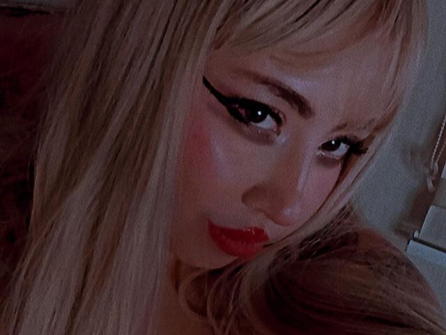 Yuri69, con i capelli biondi, un ombretto nero intenso e il rossetto rosso, guarda seducentemente la fotocamera con le labbra rosse leggermente dischiuse. Il suo viso è vicino alla macchina fotografica, con una luce soffusa..