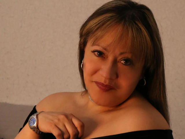 ChristinMoore a les cheveux bruns, un léger sourire, et porte un haut noir à une épaule, qui dévoile ses seins. Elle porte une montre en argent et de petites boucles d'oreilles cercles..
