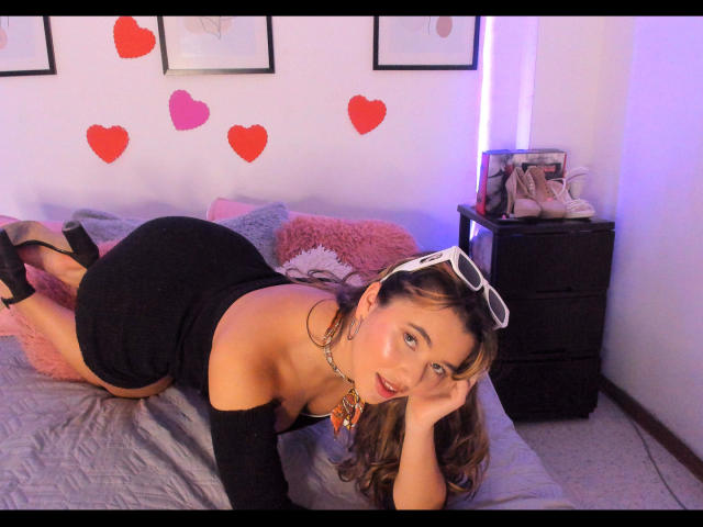 KatherineSummer, laying on a bed, black off-shoulder dress, showing her ass, wearing black high heels, white sunglasses on head, necklace, red lipstick, brown hair, wall with heart decals.