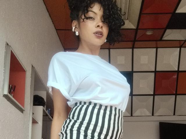 Anna, con i capelli neri ricci, indossa una t-shirt bianca e pantaloni a vita alta a righe nere e bianche, mostrando il seno sotto la t-shirt, con rossetto rosa e orecchini di perle..